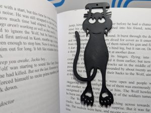 A black 3D-printed plastic bookmark shaped like a flattened, cartoonish cat resting between the pages of an open book.